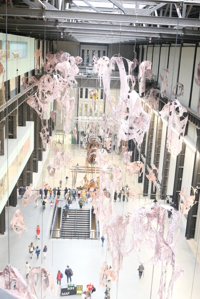 現代美術館の内部、天井から吊るされたアート作品があり、来場者が階段を進んでいる様子。