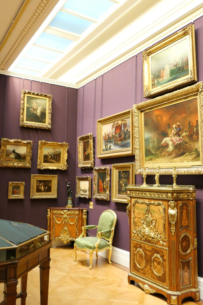 美術館の内部、紫色の壁に多数の絵画が掛けられている。左側には木製の家具と彫像がある。